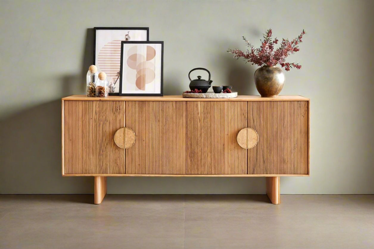 A large sideboard in a natural wood finish with four doors and circular handles, displayed in a room with decorative items and framed artworks on top.