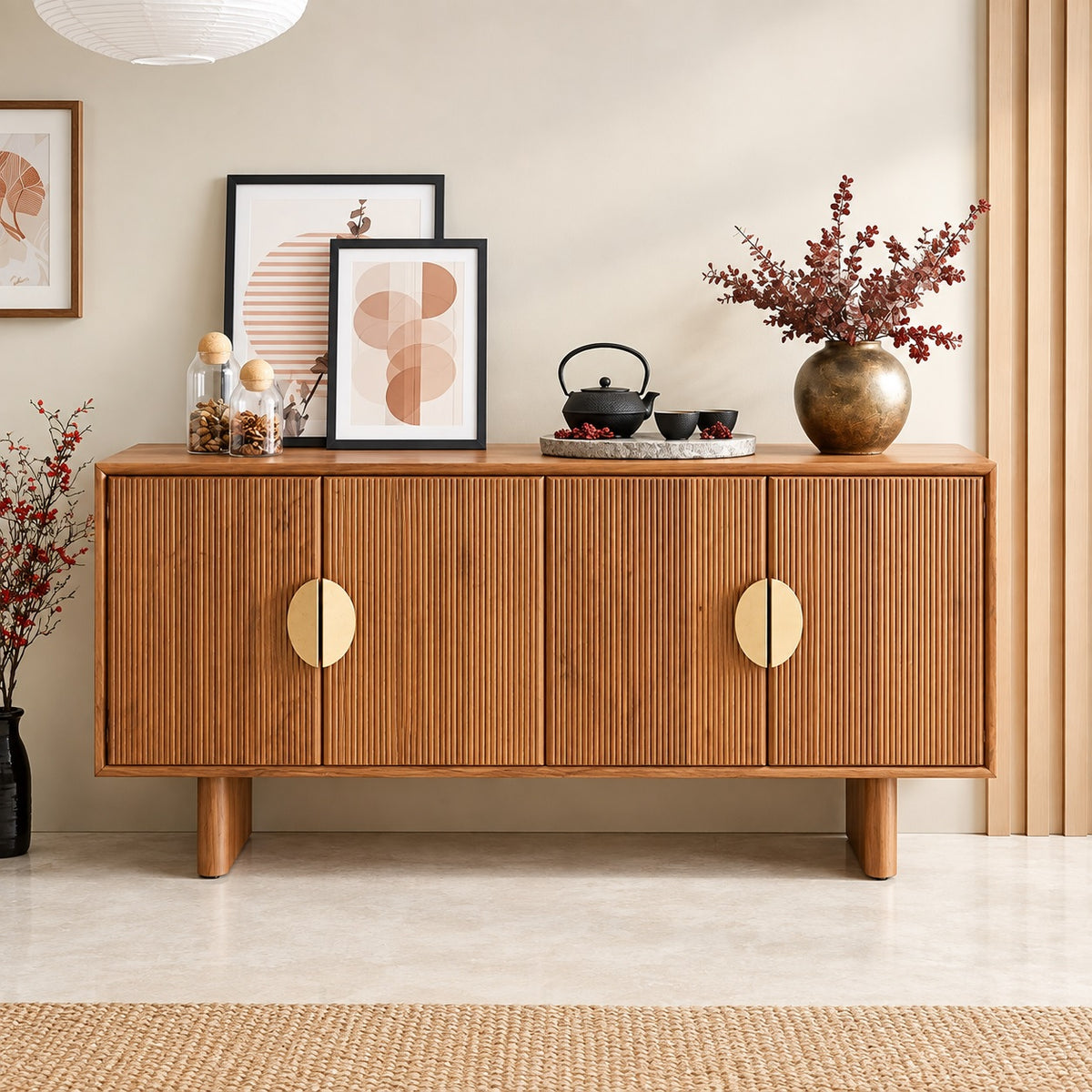A mid-century modern wooden sideboard cabinet with vertical fluted panels and oval brass handles, styled in a warm beige room with framed art, a teapot set, and a vase of branches.