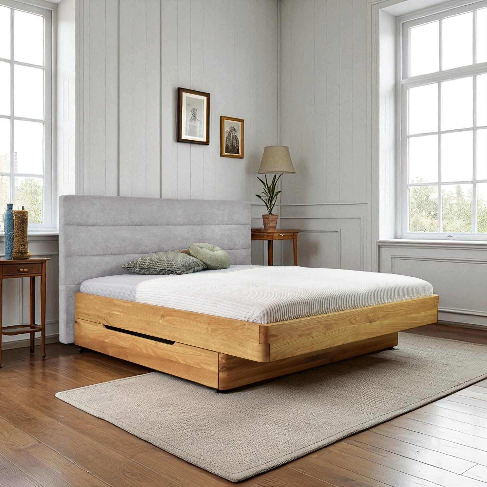 Wooden bed with gray upholstered headboard in a room with large windows and light-colored walls.