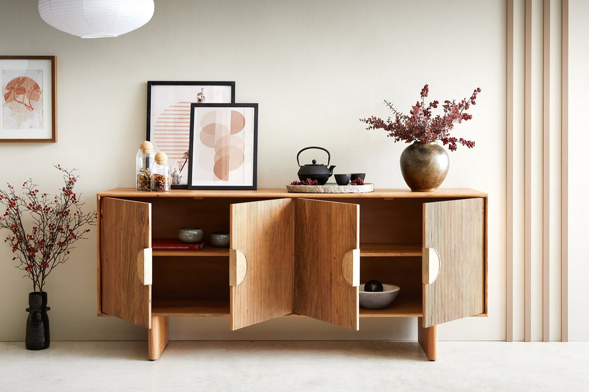 A natural finish large sideboard made of solid wood, with four doors and a flat top, placed in a room with decorative items on top and against the wall.