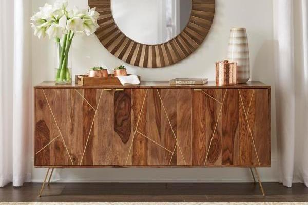 A rustic teak finish sideboard with three doors and iron legs, placed against a backdrop of a living room with decorative items on top.
